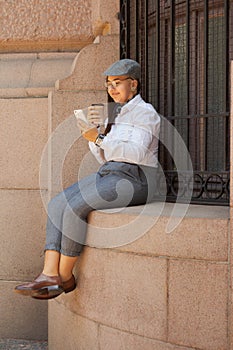 Woman with cup of coffee watching photos on mobile smart phone sitting in old building window. Technology, social