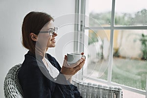 Woman with a cup of drink sits in an armchair near the window at home interior