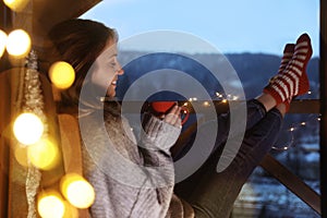 Woman with cup of hot beverage and Christmas lights resting on balcony