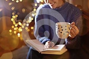 Woman with cup of hot beverage reading book at home in winter evening