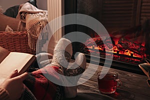 Woman with cup of tea reading book near fireplace at home, closeup