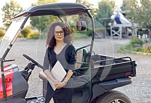 Woman with curly hair in black dress holding in hands a laptope, poses near vehicle and smiling. Small bussines content