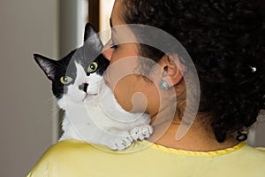 Woman with curly hair holding and kissing her domestic black and white kitten with green eyes on the shoulder. Concept