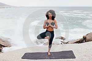 Woman with curly hair practicing Tree Pose by seaside. Young female with closed eyes doing Vrksasana at evening