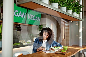 Woman with curly hair seats at vegetarian cafe or restaurant with green smoothie and plant meal on table, looking