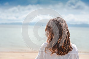 A woman with curly hair stands with her back in the wind on the beach. Sea and travel, hair care