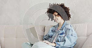 Woman with curly hair writing message in social media on laptop sitting on sofa.