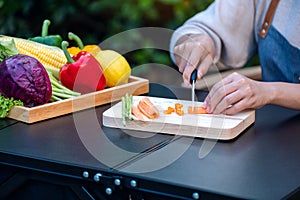 A woman cutting and chopping carrot by knife on wooden board with mixed vegetables in a tray