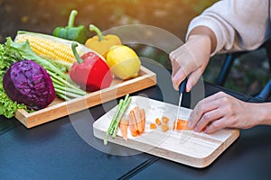 A woman cutting and chopping carrot by knife on wooden board with mixed vegetables in a tray