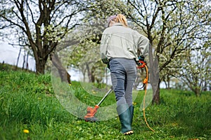 Woman is cutting grass by string trimmer in orchard