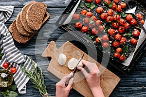 Woman cutting mozzarella cheese for sandwiches with herbs and baked tomatoes on dark