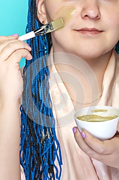 A woman with dark blue afro braids applies a mask of green clay to her face with a brush on a blue background part of her face