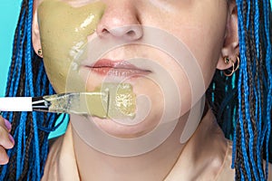 woman with dark blue afro braids applies a mask of green clay to her face with a brush on a light blue background