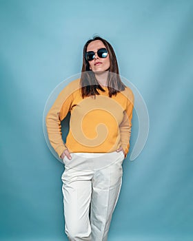 Woman in dark glasses, yellow sweater stands hands in pockets on blue background