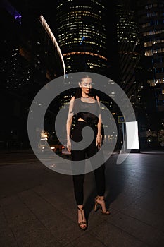 woman with dark hair in elegant dress and accessories walks around the night city with skyscrapers in the background