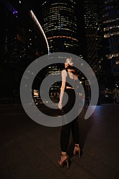 woman with dark hair in elegant dress and accessories walks around the night city with skyscrapers in the background