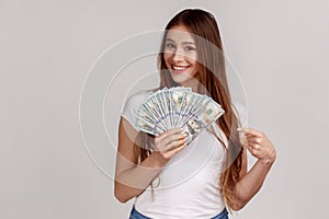 Woman with dark hair holding fan of dollars and pointing at banknotes, looking at camera.