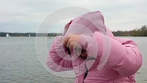 Woman with dark hair zips her pink coat puts on hood and scarf. Cold autumn day during pandemic.