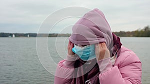 Woman with dark hair zips her pink coat puts on hood and scarf and mask. Cold autumn day during pandemic.