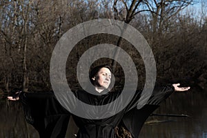 Woman with dark long hair in black robes in front of the lake. Magic and Witchcraft. Back to Nature concept