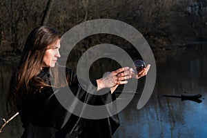Woman with dark long hair in black robes is holding polished Clear Quartz Sphere in her hands in front of the lake