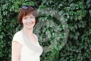 Woman with dark red hair stands and smiles in front of a wall covered in leaves