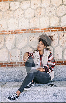 Woman with dark skin and afro hair sitting on a stone bench, smiles while she is thinking something to write in her notebook