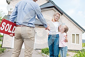 woman with daughter buying new house and shaking hand of male realtor with sold sign