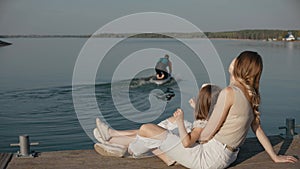 Woman with daughter sit on a pier look at dad surfing on a lake at the sunset