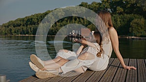 Woman with daughter sit on a pier looking at lake smiling at the sunset