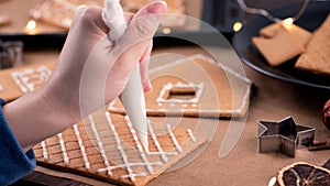 Woman is decorating gingerbread cookies house with white frosting icing cream topping on wooden table background, baking paper in