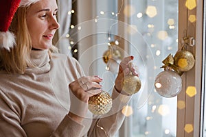 woman decorating window using golden christmas balls and lights bokeh. blond wearing red santa hat