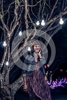 Woman among decorative outdoor string lights hanging on tree in the park at night time. Bali island, Indonesia.