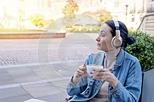 woman in denim coat and headphones drinking coffee and listening to music or audiobook in the cafe.