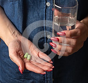 a woman in a denim shirt holds a glass of water in one hand and in the other hand two tablets on her palm. illness, headache,