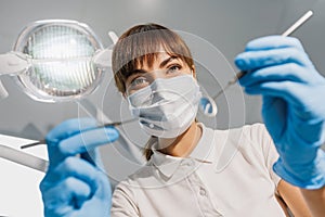 Woman dentist in blue face mask and gloves examine teeth with probe and mirror. View from patient seat to doctor