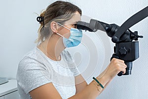 Woman dentist in medical mask looking through binocular of dental microscope and holding handles of innovative device.