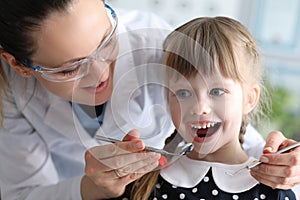Woman dentist in protective glasses examines teeth of little girl patient with metal tools portrait