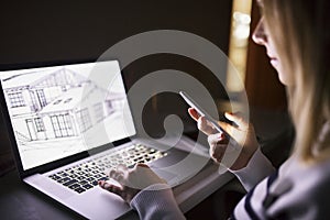 Woman at desk, holding smartphone, working on laptop at night.