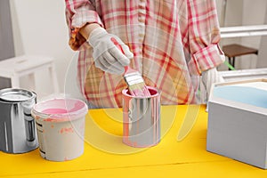 Woman dipping brush into can of pink paint at yellow wooden table indoors, closeup