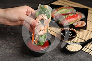 Woman dipping roll wrapped in rice paper into sauce at grey table, closeup