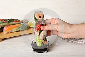 Woman dipping roll wrapped in rice paper into sauce at white wooden table, closeup
