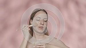 A woman dips a makeup brush into a jar of powder, blush and applies cosmetics to her face. Portrait of a seminude woman