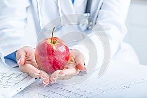 Woman doctor in ambulance holds red apple in hands. On desk laptop and EKG diagram