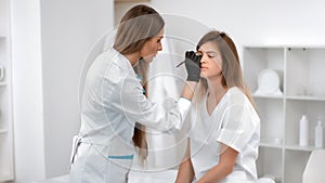 Woman doctor cosmetologist correcting improving shape and color of eyebrows client at beauty salon