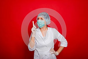 A woman doctor, dressed in a white coat, mask, medical cap and rubber gloves, stands on a red background and holds his index finge photo