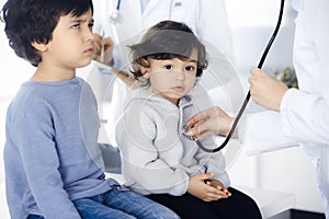 Woman-doctor examining a child patient by stethoscope. Cute arab toddler and his brother at physician appointment