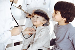 Woman-doctor examining a child patient by stethoscope. Cute arab toddler and his brother at physician appointment