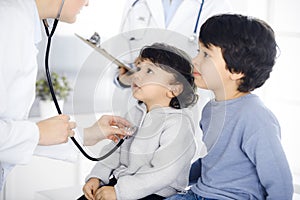 Woman-doctor examining a child patient by stethoscope. Cute arab toddler and his brother at physician appointment