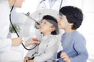 Woman-doctor examining a child patient by stethoscope. Cute arab toddler and his brother at physician appointment